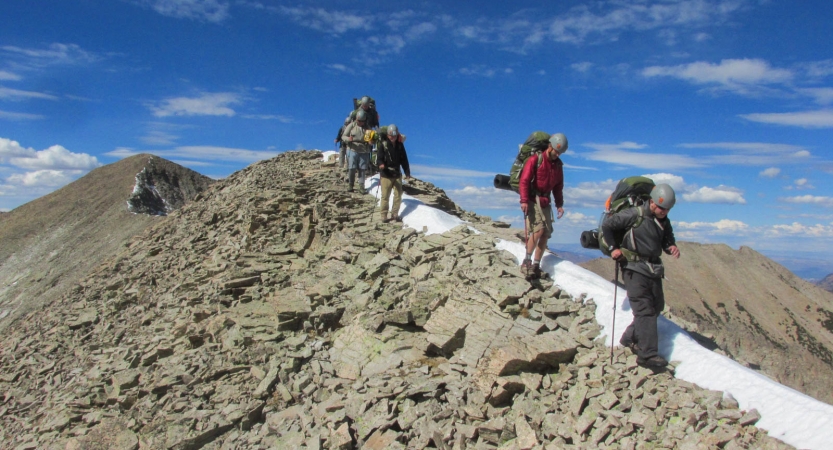 A group of people carrying backpacks and wearing safety gear hike along a rocky ridge with blue skies above.